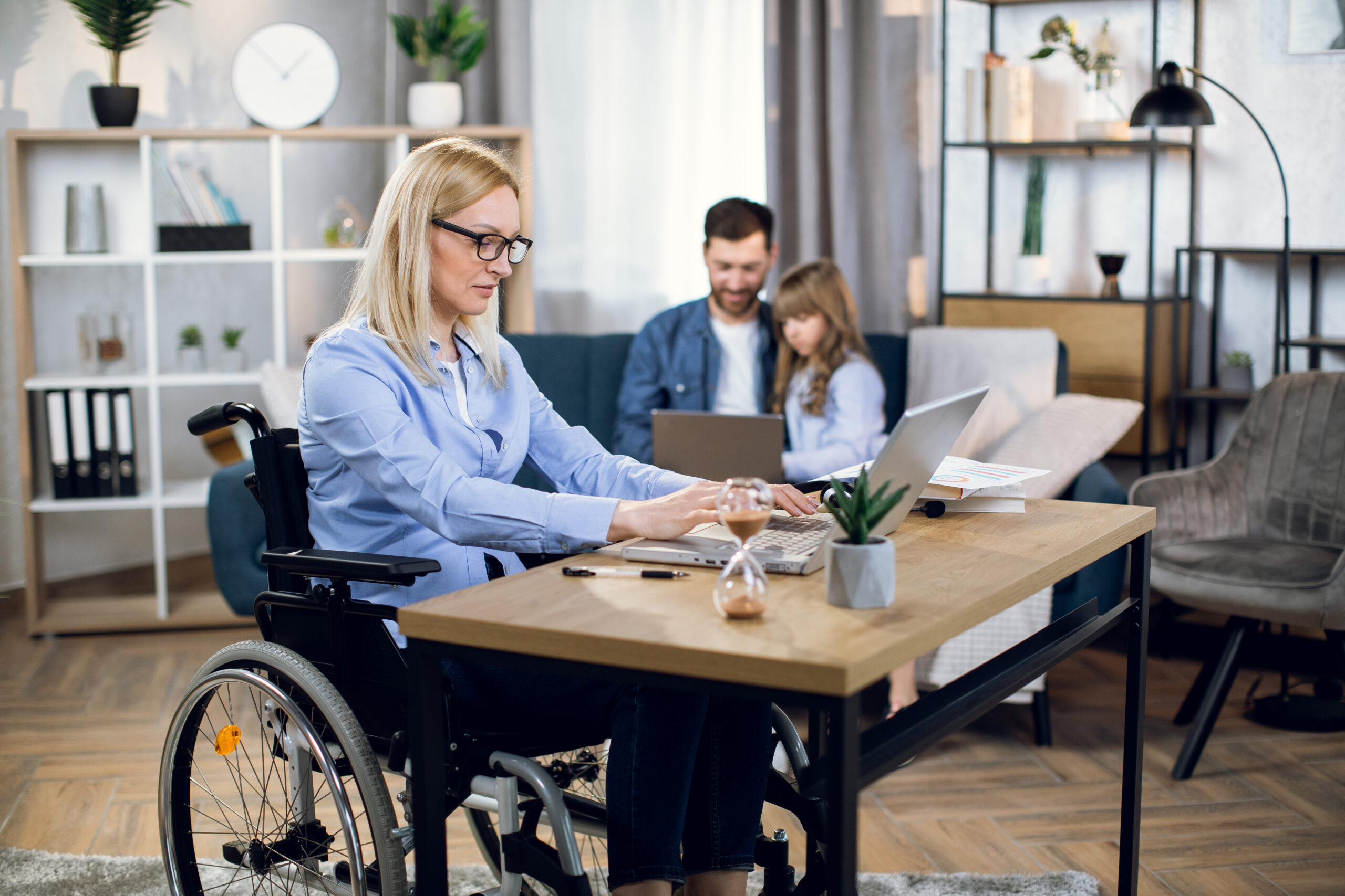 Woman with special needs working on laptop Side view of attractive woman with special needs sitting at table and working on wireless laptop. Husband with daughter playing on computer. Concept of disability, freelance and family support.