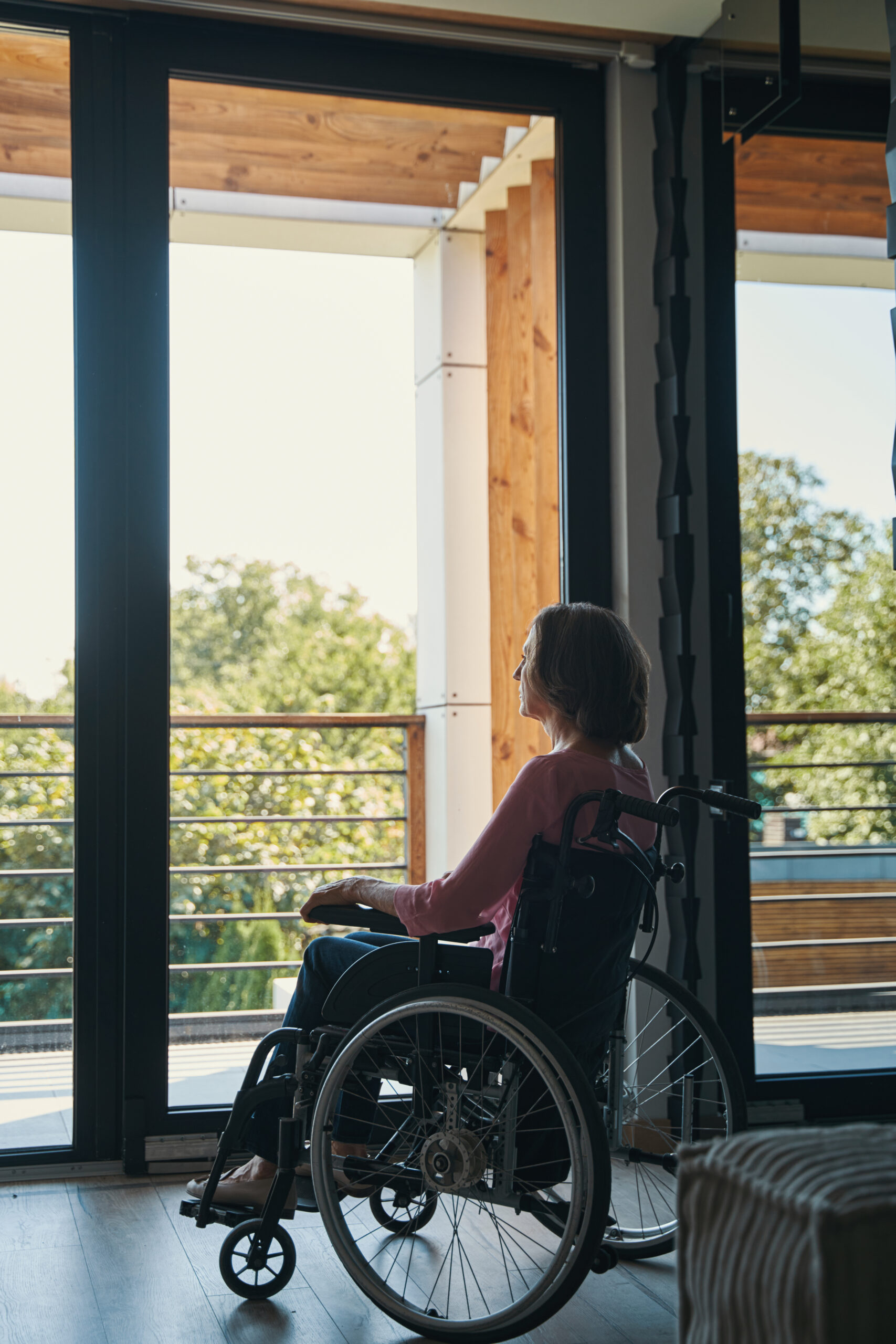 Disabled woman in wheelchair staring at window Senior female with disability sitting in manual wheelchair and looking out of the window